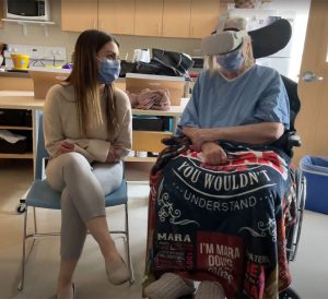 Older woman, sitting on a wheelchair, wearing a virtual reality headset. A lady is sitting beside her.